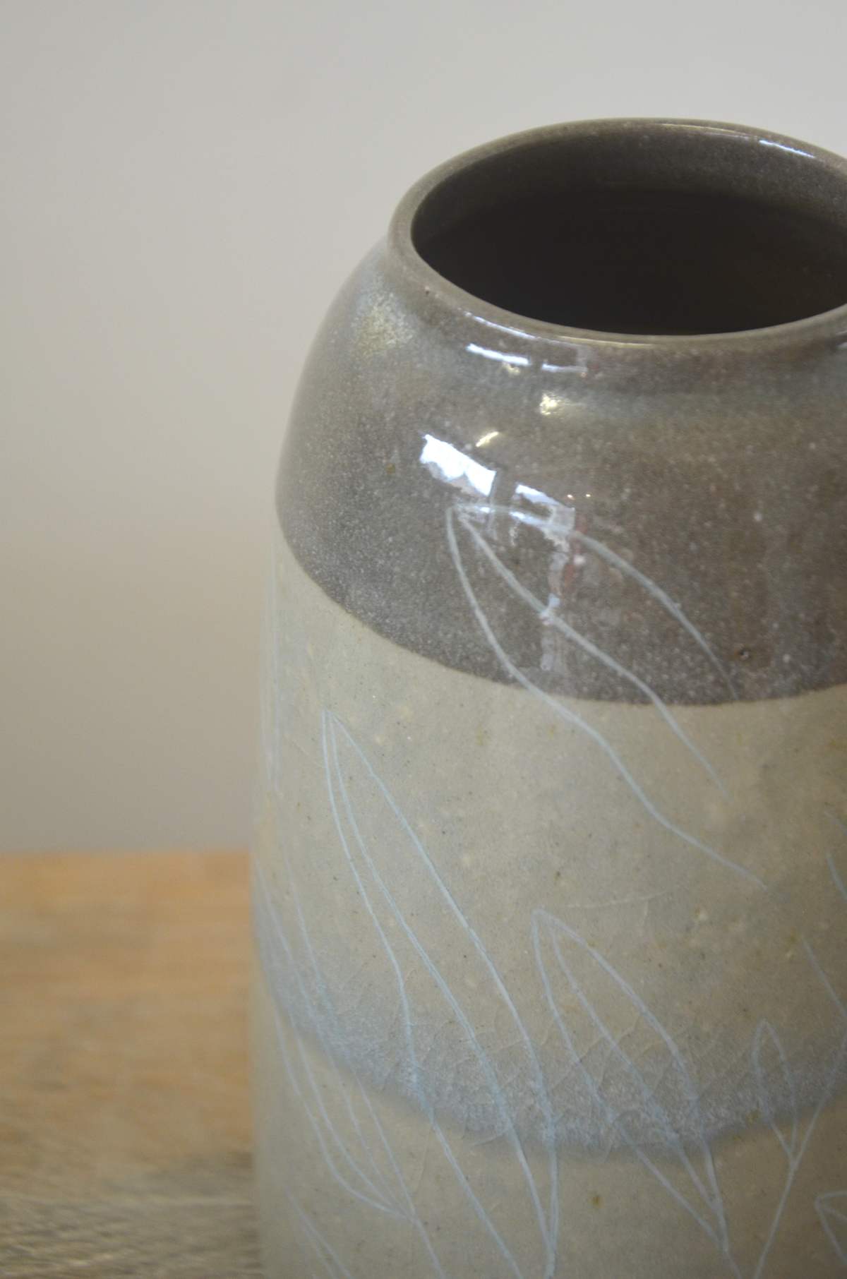 Détail du col gris et brillant d'un vase, feuillages clairs sur la paroi.