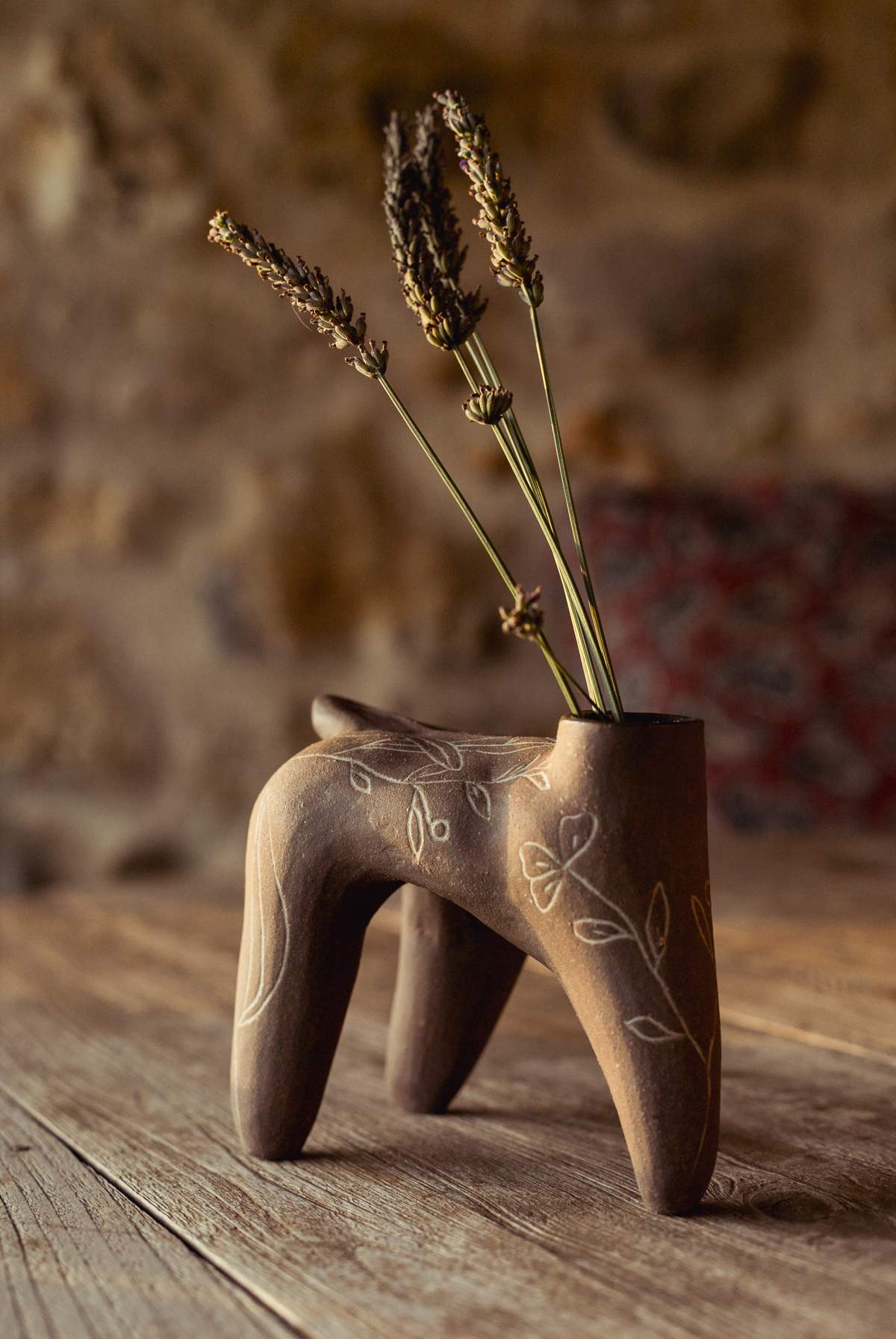 Pièce brune à quatre pieds, feuillages incisés, tenant des épis de graminées séchées, sur une table en bois devant un mur de pierre.